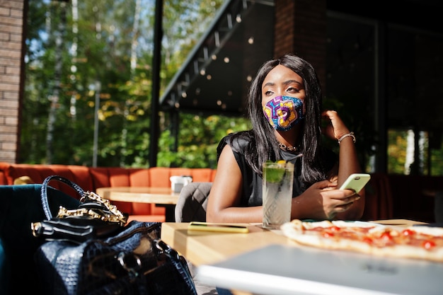 Photo gratuite une femme afro-américaine à la mode porte un masque facial fait à la main assis au restaurant regarde son téléphone portable tout en mangeant de la pizza nouvelle vie normale