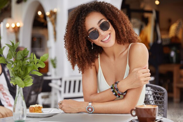 Femme afro-américaine à la mode souriante heureuse dans des lunettes de soleil à la mode, boit du café ou du café au lait, mange un gâteau sucré, aime le temps libre au café