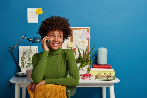 Une femme afro-américaine parle via un téléphone intelligent, travaille au bureau au bureau à domicile, a une expression de visage joyeuse