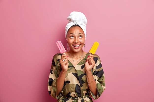 Une femme afro-américaine à la peau foncée positive concentrée au-dessus porte une robe de chambre et une serviette enveloppée de blanc sur la tête tient deux délicieuses glaces sur des bâtons posés contre un mur rose. Style de maison