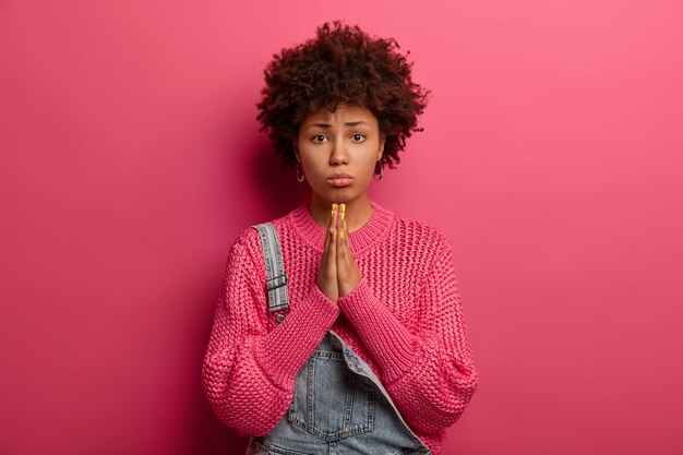 Une femme afro-américaine à la peau sombre et regrettable demande pardon, se sent coupable, porte un sac à main, porte un chandail tricoté, isolé sur un mur rose, implore les parents pour de l'argent, semble innocent