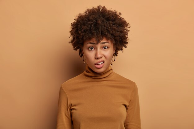 Une femme afro-américaine perplexe a déplu à l'expression, fronce les sourcils, regarde avec dégoût quelque chose de désagréable, vêtue de vêtements décontractés, isolée sur un mur marron. Émotions et réaction