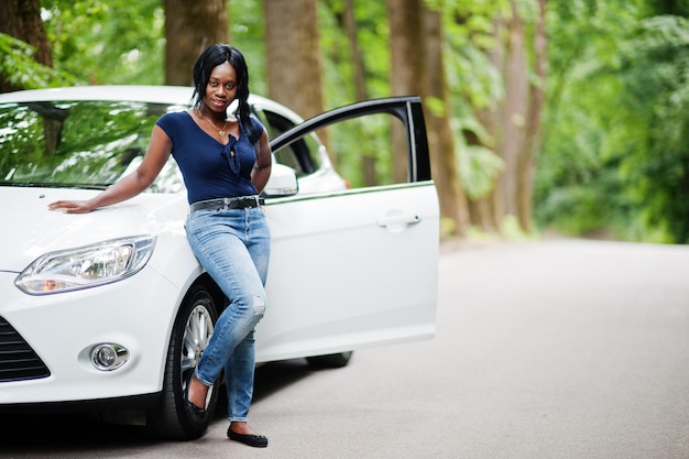 Femme afro-américaine posée contre une voiture blanche sur une route forestière