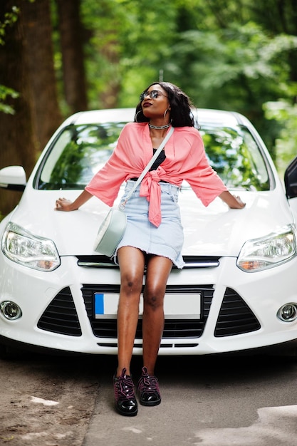 Femme afro-américaine posée contre une voiture blanche sur une route forestière