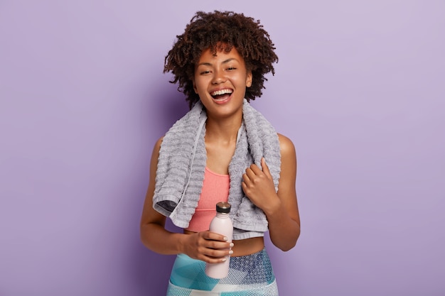 Femme afro-américaine positive insouciante vêtue de vêtements de sport, tient une bouteille d'eau fraîche, heureuse d'être en bonne santé, a une formation régulière, porte une serviette douce sur le cou