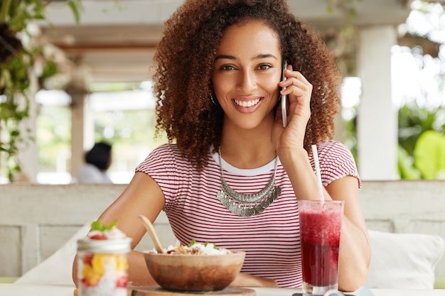 Une femme afro-américaine positive a un large sourire brillant, communique par téléphone portable pendant la pause dîner dans un café exotique, a une conversation agréable avec ses proches, partage ses impressions sur les vacances