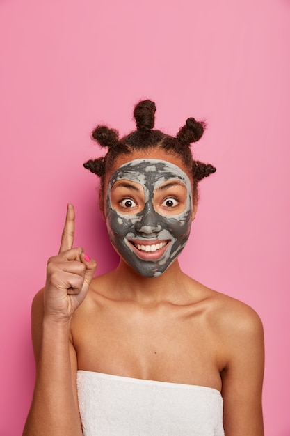 Une femme afro-américaine positive pointe ci-dessus avec l'index, se soucie de la peau, applique un masque d'argile, fait la publicité d'un beau produit de beauté, se soucie de l'apparence, a l'air jeune et belle, pose à l'intérieur