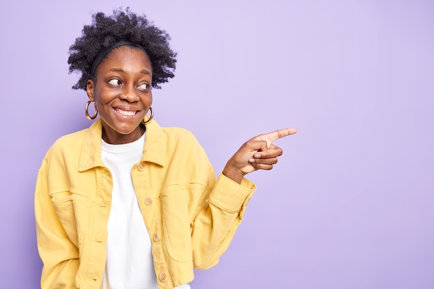 Une femme afro-américaine ravie aux cheveux noirs et bouclés sourit agréablement, indique à droite l'espace de copie