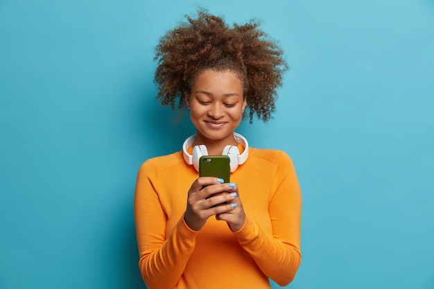 Une femme afro-américaine ravie d'être accro aux réseaux sociaux et aux technologies modernes détient des messages texte de types cellulaires porte des écouteurs stéréo autour du cou habillés de vêtements décontractés