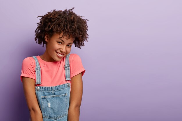 Une femme afro-américaine ravie a une expression de visage joyeuse et timide, exprime des émotions positives, porte un t-shirt rose et un sarafan en denim, des modèles sur un mur violet, copiez l'espace de côté