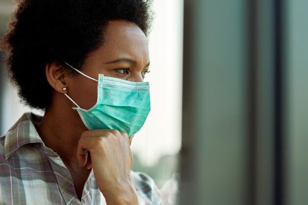 Femme afro-américaine réfléchie portant un masque protecteur et regardant par la fenêtre