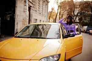 Photo gratuite femme afro-américaine en robe violette et casquette posée sur une voiture jaune