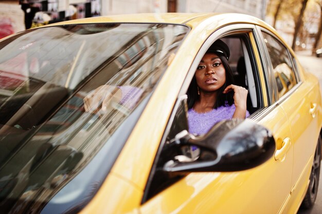 Femme afro-américaine en robe violette et casquette posée sur une voiture jaune