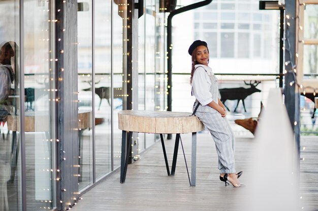 Femme afro-américaine en salopette et béret posé sur une terrasse extérieure avec guirlande de décorations de noël