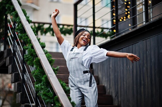 Femme afro-américaine en salopette et béret posé sur une terrasse extérieure avec guirlande de décorations de noël