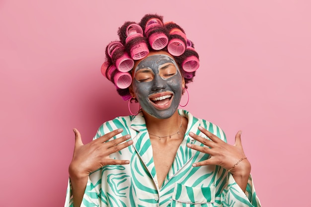 une femme afro-américaine satisfaite sourit largement apprécie les traitements de soins de la peau applique un masque d'argile nourrissant sur le visage fait une coiffure frisée porte une robe de chambre en soie isolée sur un mur rose