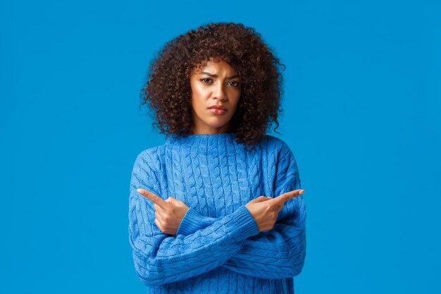 Femme afro-américaine sceptique et incertaine, indécise et sérieuse avec une coupe de cheveux afro, fronçant les sourcils et incertaine, pointant les doigts sur le côté, à gauche et à droite sur fond bleu.