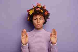 Photo gratuite une femme afro-américaine sérieuse fronce les sourcils et montre un geste d'arrêt, porte un pull tricoté chaud, garde les paumes tendues à la caméra, a des feuilles d'automne coincées dans les cheveux bouclés. saison, langage corporel