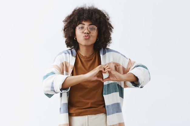 Photo gratuite femme afro-américaine sociable élégante et moderne dans des verres et un cardigan rayé élégant montrant le signe du cœur sur la poitrine se pliant les lèvres en baiser ou mwah exprimant la sympathie et l'affection