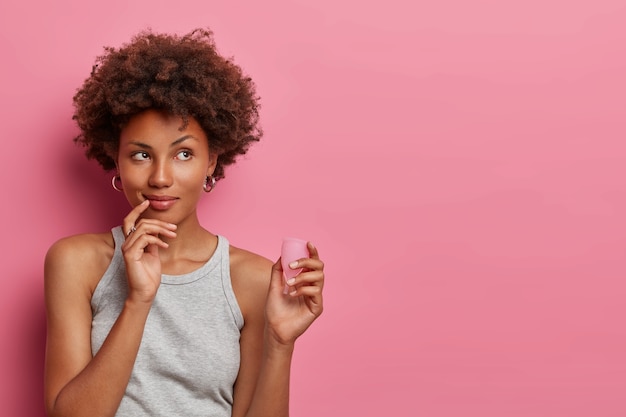 Une femme afro-américaine songeuse détient un produit menstruel efficace et abordable, une bonne protection contre les fuites, choisit une coupe menstruelle réutilisable, réfléchit à son utilisation, un espace vide vide sur un mur rose