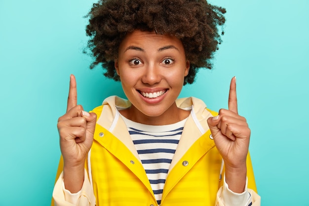 Femme afro-américaine souriante avec une expression heureuse, montre la direction ci-dessus, porte un pull rayé et un imperméable jaune, isolé sur fond bleu.