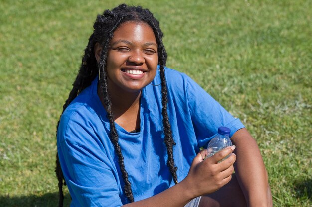 Femme afro-américaine souriante se reposant après l'entraînement. Femme potelée en T-shirt avec nez percé assis sur l'herbe, tenant de l'eau en bouteille. Sport, concept positif pour le corps