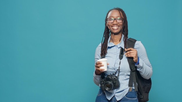 Femme afro-américaine tenant une tasse de café et un sac à dos avant de se rendre en vacances pour prendre des photos. Partir en vacances avec appareil photo et matériel photo, prendre des photos.