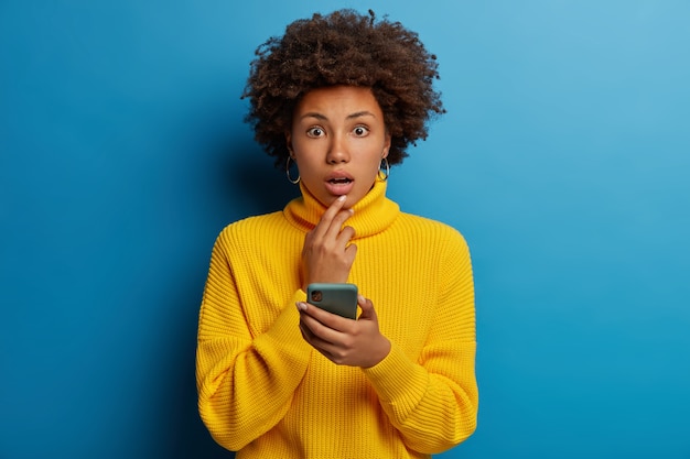 Une femme afro choquée reçoit une offre de réduction d'achat inattendue et tient un téléphone portable moderne en mains