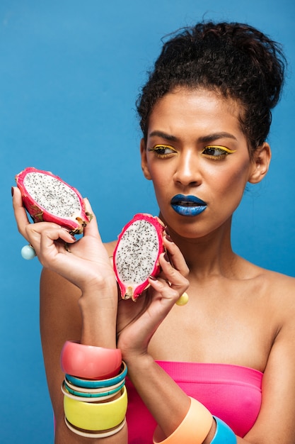 Femme afro fascinante avec des cosmétiques colorés sur le visage à la recherche sur les fruits du dragon juteux coupés en deux tenant dans les deux mains isolées, sur le mur bleu