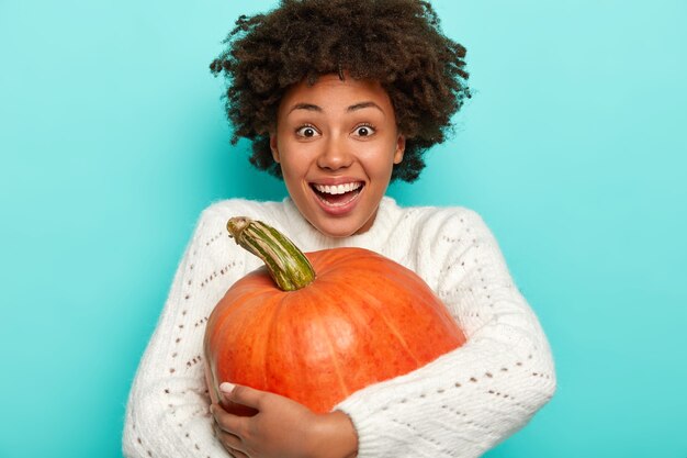 Femme afro ravie embrasse la grosse courge, sourit largement, heureuse de récolter les cultures d'automne, porte un pull blanc tricoté