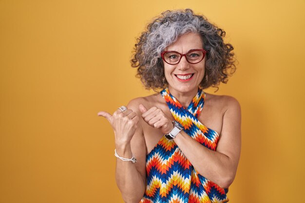 Femme d'âge moyen aux cheveux gris debout sur fond jaune pointant vers l'arrière derrière avec la main et les pouces vers le haut, souriant confiant