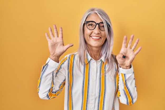 Femme d'âge moyen aux cheveux gris debout sur fond jaune portant des lunettes montrant et pointant vers le haut avec les doigts numéro neuf tout en souriant confiant et heureux