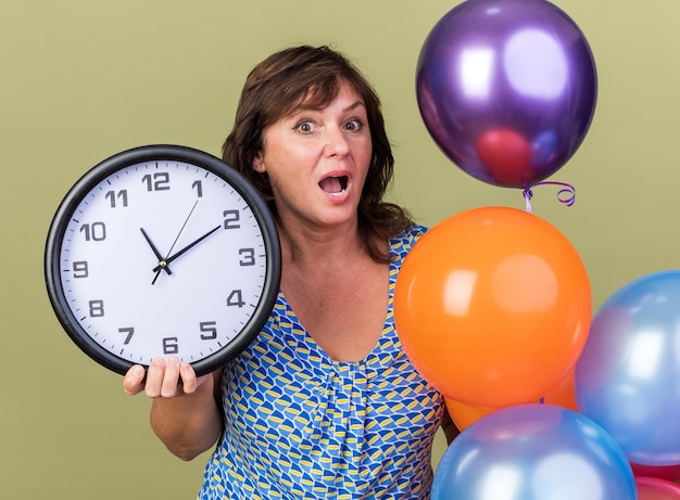 Photo gratuite femme d'âge moyen avec un bouquet de ballons colorés tenant une horloge murale étonnée et surprise