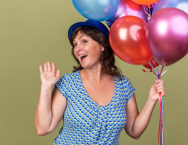 Femme D'âge Moyen En Chapeau De Fête Avec Bouquet De Ballons Colorés Heureux Et Excité Souriant Joyeusement Avec Le Bras Levé Célébrant La Fête D'anniversaire Debout Sur Un Mur Vert