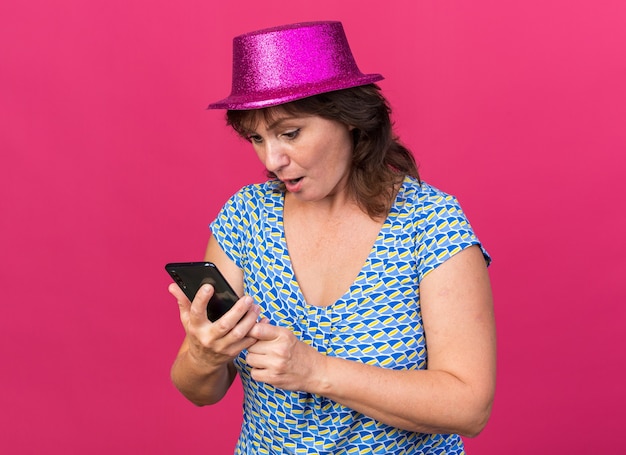 Femme d'âge moyen en chapeau de fête regardant l'écran de son smartphone surpris