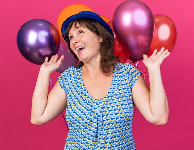 Femme d'âge moyen en chapeau de fête tenant des ballons colorés heureux et excité souriant joyeusement