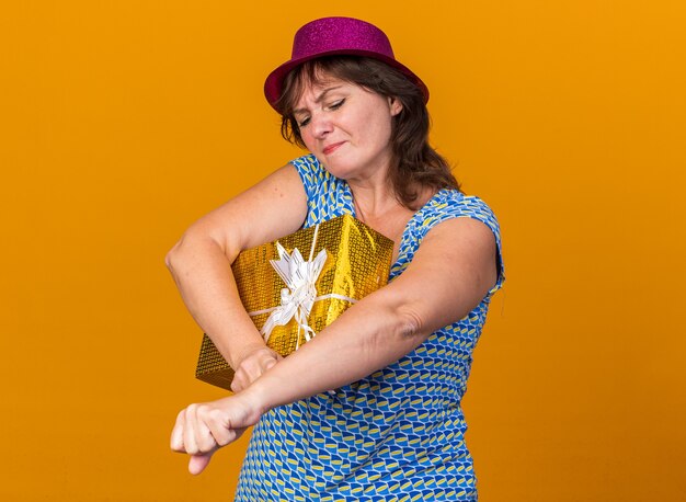 Femme d'âge moyen en chapeau de fête tenant un cadeau à la fois confus et mécontent de célébrer la fête d'anniversaire debout sur un mur orange