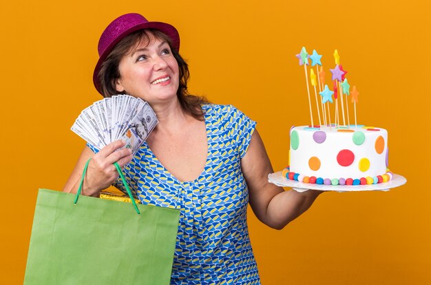 Femme d'âge moyen en chapeau de fête tenant un sac en papier avec des cadeaux tenant un gâteau d'anniversaire et de l'argent heureux et heureux souriant joyeusement célébrant la fête d'anniversaire debout sur un mur orange