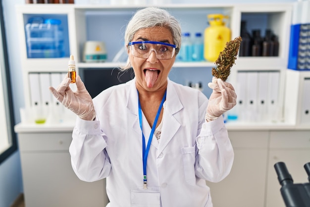 Femme d'âge moyen avec des cheveux gris faisant l'extraction d'huile de mauvaises herbes au laboratoire sticking tongue out heureux avec une drôle d'expression