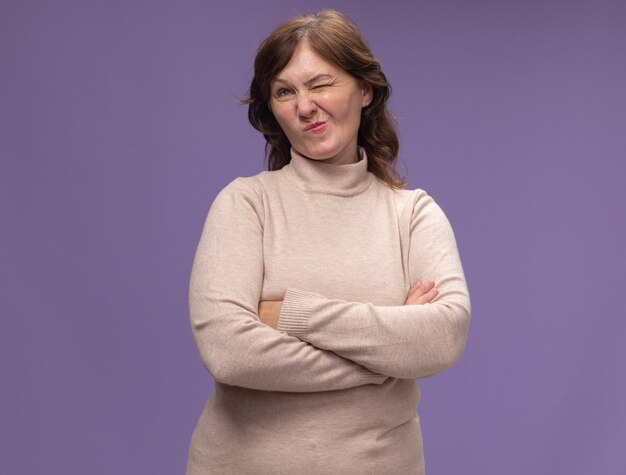 Femme d'âge moyen en col roulé beige à côté avec expression sceptique avec les bras croisés debout sur le mur violet