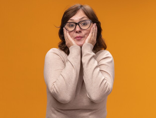 Femme d'âge moyen en col roulé beige portant des lunettes heureux et surpris debout sur un mur orange