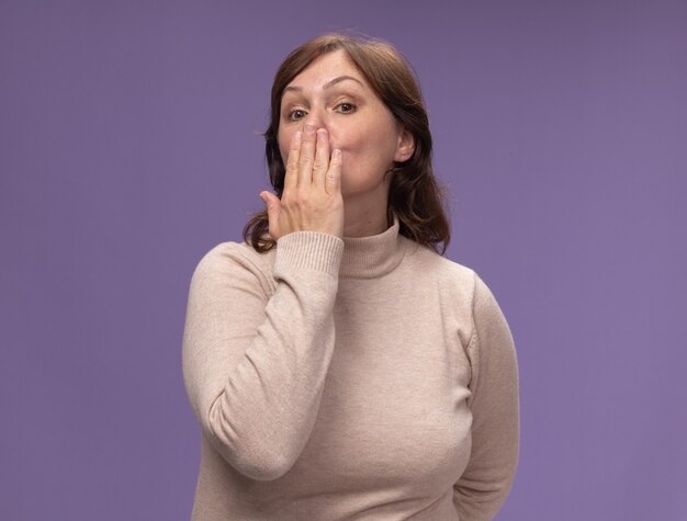 Femme d'âge moyen en col roulé beige soufflant un baiser debout sur un mur violet