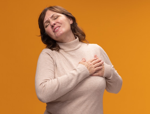 Femme d'âge moyen en col roulé beige tenant la main sur sa poitrine se sentant reconnaissante avec les yeux fermés debout sur le mur orange