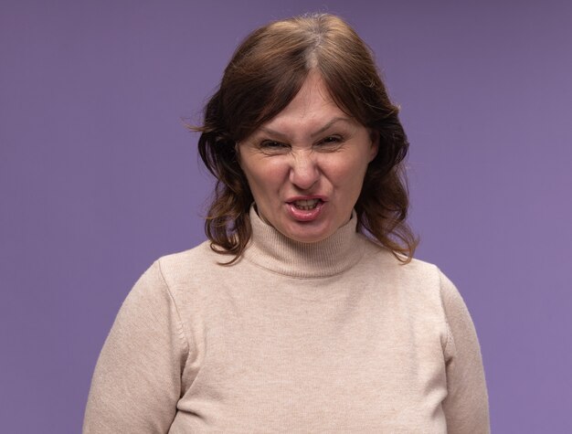 Femme d'âge moyen en col roulé beige avec visage en colère debout sur un mur violet
