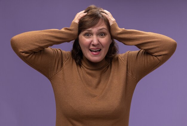 Femme d'âge moyen en col roulé marron confondu avec les mains sur sa tête pour erreur debout sur un mur violet