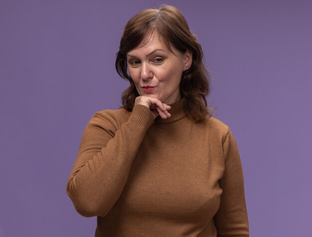 Femme d'âge moyen en col roulé marron à côté avec la main sur le menton pensant positif debout sur mur violet