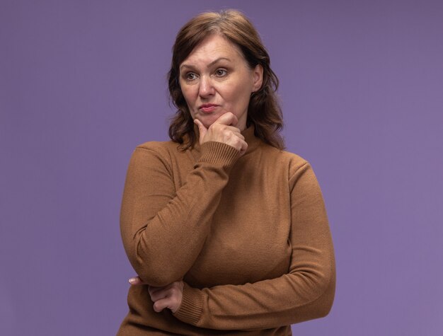 Femme d'âge moyen en col roulé marron à côté avec visage sérieux avec la main sur le menton pensant debout sur le mur violet
