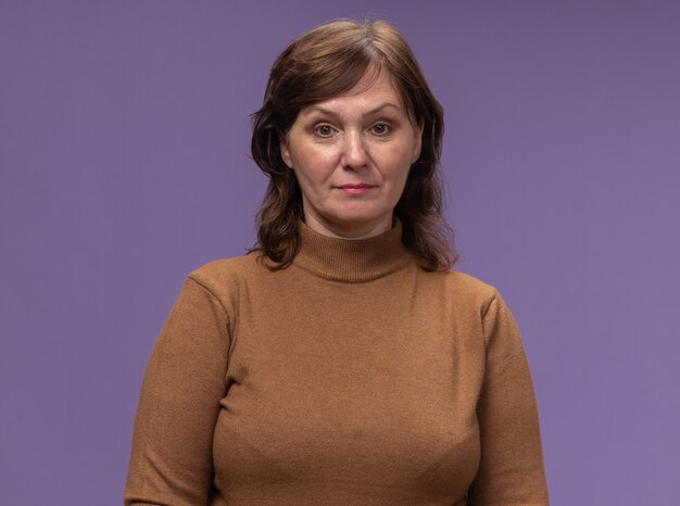 Femme d'âge moyen en col roulé marron avec expression confiante debout sur un mur violet