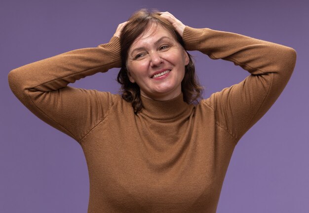 Femme d'âge moyen en col roulé marron heureux et heureux à la recherche de côté avec les mains sur sa tête debout sur un mur violet