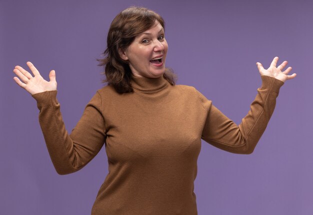 Femme d'âge moyen en col roulé marron heureux et positif large ouverture mains debout sur mur violet